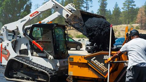 Adult in bobcat machine dumping asphalt into paver machine.