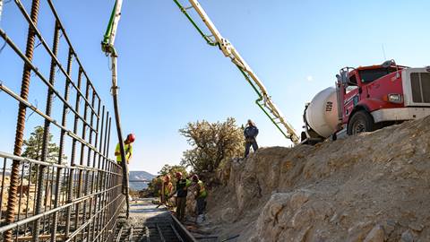 A white and red truck pouring concrete into a metal framed structure as five construction workers in yellow vests and hard hats assist where the foundation is poured.