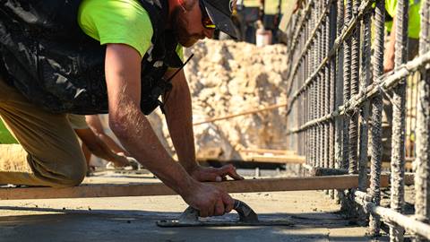 A employee in black hat, glasses, black vest, yellow shirt, and khaki pants kneeling on a wood plant smoothing out concrete next to a metal wire frame.