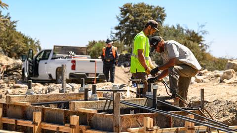 A worker in black clothing and orange vest oversees the work efforts of two construction workers, one in yellow shirt and the other in grey shirt, working on a metal and wood frame during a project on the mountain during summer.