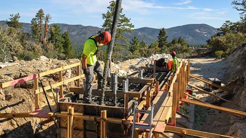 A worker in a red hard hat and yellow shirt pouring concrete into a metal and wood frame on the ski hill, during a blue sky summer day, and two other workers assist in the background.