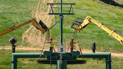 Two employees in red hard hats and yellow with orange vests assist two forklifts with a chair five chairlift project during summer at the base of Bear Mountain.