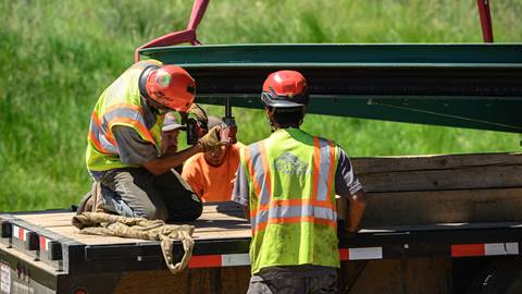 A crew of four construction workers in yellow and orange shirts on the back of a flatbed truck while one of the members with a drill works on a green chairlift part.