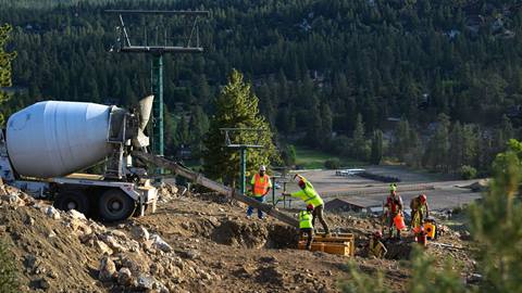 A concrete truck near a chairlift line on the Bear Mountain ski hill, during summer, while six construction crew members shovel the concrete mix into a dirt trench with a wooden frame.