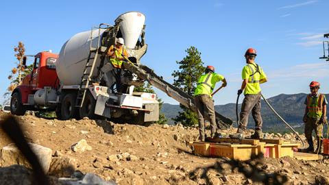 Four construction crew memebers in dark pants, yellow vests, and hard hats shovel concrete from a truck into a wooden frame in dirt on the ski hill during a blue sky summer day.