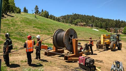 Four construction workers in black apparel, hard hats, and orange vests at the base area of a ski hill during summer working with a large metal lift cable with generators and a yellow telehandler forklift in the foreground.