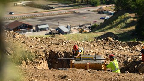 Two construction members in red hard hats and yellow vests, level out concrete that is sitting in a wooden frame in a dirt trench on the ski hill with the Bear Mountain parking lot in the distance during summer.