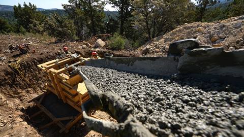 A close up image of gray concrete being poured into a wooden frame in a dirt trench on the slopes of a ski hill during a blue sky sunny summer day.