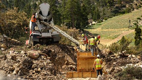 Five construction workers, in hard hats and orange yellow vests, shoveling concrete from a white truck parked next to big rocks at Bear Mountain during the summer time with green pine trees and dirt ski hill in the background.