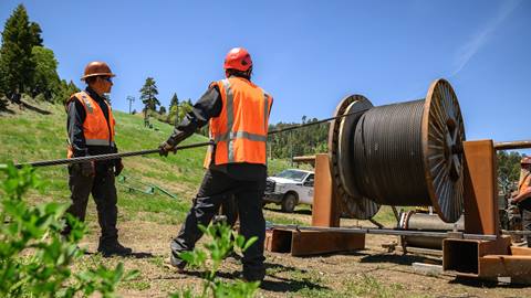 Two workers, in red hard hats with black clothing and orange vests, working with a metal cable at the base of Bear Mountain during the summer with green grass and pine trees in the fore and background.