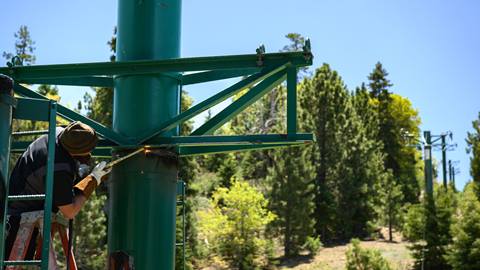 An employee in black clothing. a dark beanie, and heavy work gloves welding a piece to a green chairlift pole during a blue sky summer day.