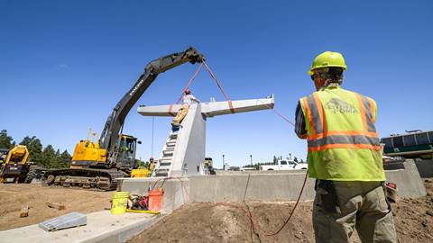 A construction worker in a yellow vest and yellow hardhat holding a red rope while workers adjust a concrete chairlift fixture in the base area of Bear Mountain during a blue sky summer day.