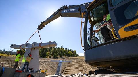 Three construction workers in yellow vests and orange and yellow hardhats working to install a concrete and metal chairlift fixture at the base of Bear Mountain while a worker operates from a excavator.