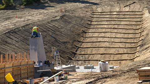 A construction worker in yellow with orange vest and black hardhat kneeling on top of a concrete fixture in a dirt trench during a new chairlift project at Bear Mountain.