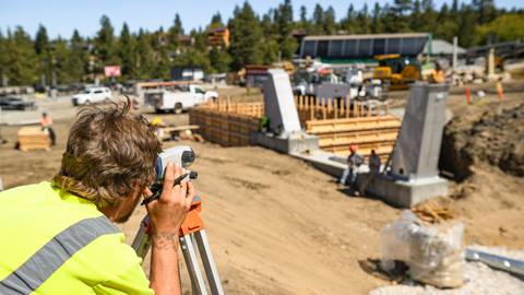 Construction site at the base of Bear Mountain during a new chairlift install project while a worker in yellow shirt holding a pen in their right hand looks through a eye measure device on a tripod.