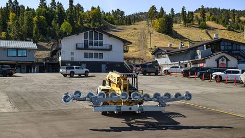 A yellow construction crane vehicle with chairlift wheels in the parking lot of Bear Mountain during a summer new chairlift project with black and white cars parked in the lot..