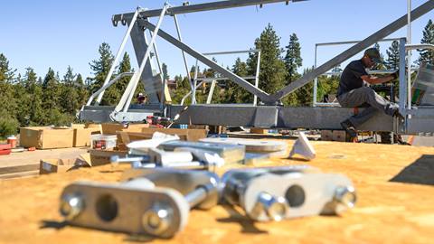 Two Big Bear Mountain Resort employees working on a metal chairlift fixture on the back of a flatbed truck during the summer with wood, cardboard boxes, and metal pieces sprawled out.