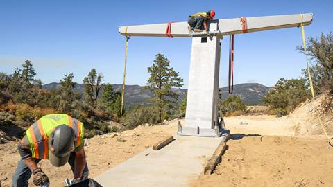 A worker in yellow and orange vest with dark hard hat bending over at a construction site on the ski hill and another worker with yellow orange vest and red hardhat on top of a chairlift fixture during a blue sky summer day.