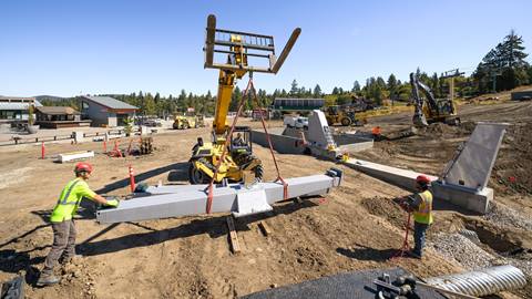 Two construction workers in red hardhats and yellow vests supporting a crane lift operator with a metal chairlift fixture at the base of Bear Mountain during summer.