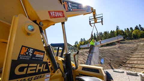A yellow construction crane suspending and holding a metal flat chairlift piece while a worker in dark pants, yellow vest, and red hardhat fixes the piece onto the base.