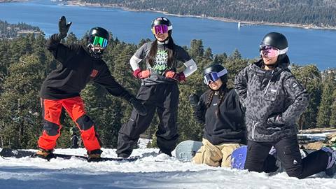 A group of four snowboarders, all wearing helmets with snow goggles, posing for a photo on a sunny winter day at Snow Summit with Big Bear Lake in the background.