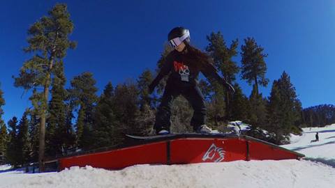 A snowboarder wearing black pants, a black long sleeve top, and a black helmet with white snow goggles, riding on top of an up-flat-down beginner rail at Bear Mountain on a sunny winter day..