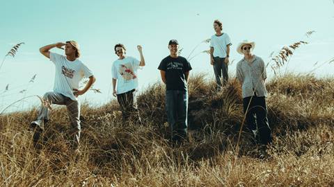 A group of five adults, wearing casual clothing, standing in a field with straw grass up to their shins on a sunny day.