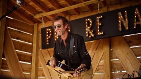 Adult in sunglasses and long sleeve black shirt playing a guitar while on a wooden stage.