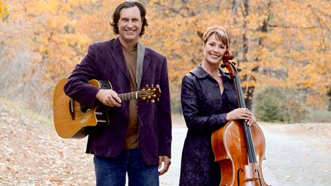 Two adults in purple coats, one holding a guitar and the other with a cello, outside during fall.