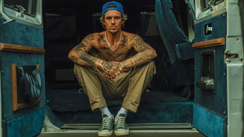 An adult in a blue backwards hat, khaki pants, Converse shoes, white high socks, and no t-shirt exposing their full torso of tattoos sitting in a van with both rear doors open.