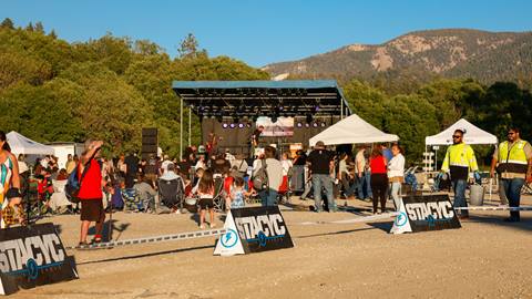 A crowd waiting for live music to begin, gathered in front of a structured stage, with eaze-up tents and a roped off STACYC track course for mini bike riders to enjoy.