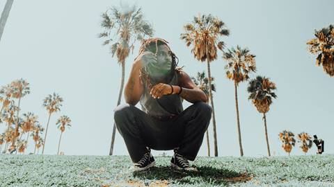 Adult in a black tank top, black jeans, and black Converse shoes squatting on grass while starring to their left surrounded by blue skies and palm trees.
