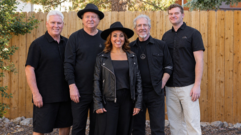 Five adults, dressed in business casual dark clothing, smiling and posing for a group band photo in front of a wooden fence with alpine trees surrounding them.
