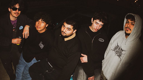 A close-up group portrait of four members of the Hellbombs band standing side by side, dressed in casual dark attire including t-shirts and hoodies, posing for a photo.