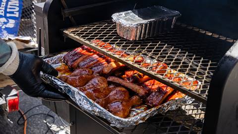 A persons hand putting a foil plate with BBQ meats onto a black grill.