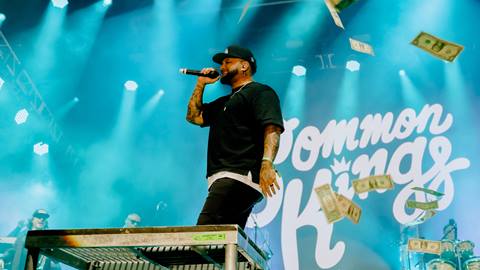 Singer for Common Kings, dressed in black hat, pants, and t-shirt, with a microphone on stage with a blue Common Kings backdrop and dollar bills falling from the sky.