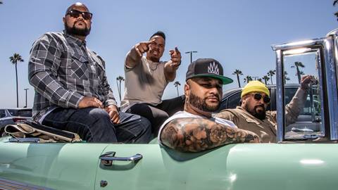 Four members of the Common Kings reggae band posing in a green old school car at the beach with palm trees in the background.