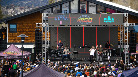 Front view of a band playing with a large crowd in front of a brown building