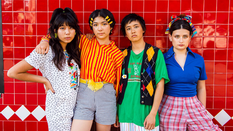 The official The Linda Lindas group photo shot, artists posing for a photo in front of a red walls