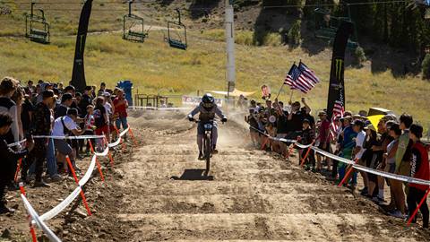 A whoop section of a mountain bike race, with crowds of fans on each side, towards the base of Snow Summit with a racers on their bike in the middle of the track.