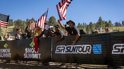A group of fans cheering from the sideline during a mountain bike race holding American flags towards the race finish line.