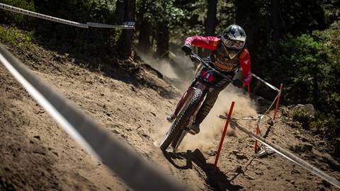 A mountain biker rider riding their bike on a dirt trail with fencing on the sides, wearing dark pants, a red jersey, and dark full face helmet.