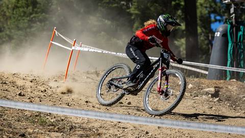 A mountain biker in a red bib with long hair, full face helmet, and goggles, racing down a dirt trail that is fenced off during a competition.