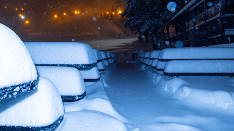 A nighttime image at Snow Summit with fresh snow on the tables, seating, and walkway in the base area while the lights turned on on the slopes.