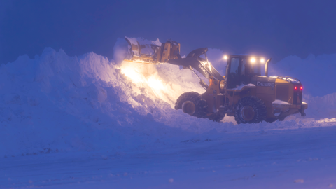 A snow plow ,with it's lights turned on in the early morning, pushing snow into a bank to clear the road.