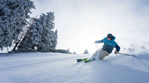 A skier, in a blue jacket, riding down an untouched powder run at Snow Valley while having a big smile on their face.