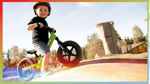 A toddler on a mini bike stopped on a small roller with the Snow Summit climbing wall in the background