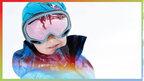 Kiddo in a blue jacket with a beanie and goggles on the ski hill smiling 