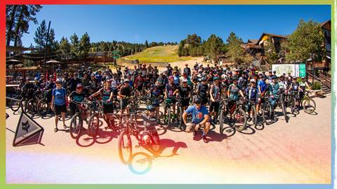 A large group of mountain bike enthusiasts posing with their bikes at the base of Summit Bike Park during a blue sky summer day.