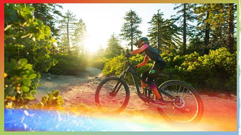 A mountain bike rider, in a matching Fox branded top and bottom mountain bike apparel set with a full face helmet on a black mountain bike, riding along a dirt trail as the sun sets during summer.